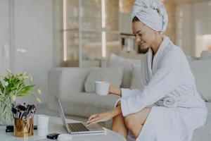 foto interna de freelancer feminina trabalha distante rom tipos domésticos no laptop vestido com roupão bebe café ou chá gosta de atmosfera doméstica navega na internet lê conselhos sobre cuidados com a pele