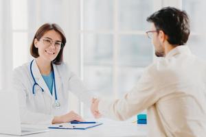 conceito de parceria, assistência, confiança e medicina. médica aperta a mão do paciente agradecido pelo bom tratamento e profissionalismo, pose na clínica, registros médicos perto da mesa branca foto