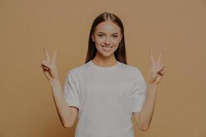 tiro horizontal de feliz otimista caucasiana jovem levanta os braços mostra gesto de paz com os dedos faz sinal de vitória diz que o número dois usa camiseta branca casual isolada sobre fundo marrom foto