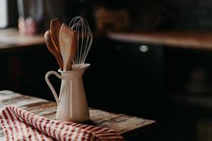 acessórios de cozinha na mesa de madeira. utensílios em jarra de cerâmica branca contra um fundo escuro. estilo rústico. louça para preparar a refeição. colheres de pau shpatula e bata foto