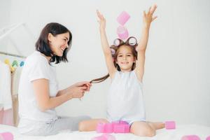 alegre menina bonita com bobes na cabeça, levanta as mãos, passa o tempo livre com a mãe, que faz penteado, estando em casa. linda mãe e filha se divertem no interior, fazem o cabelo. conceito de beleza foto