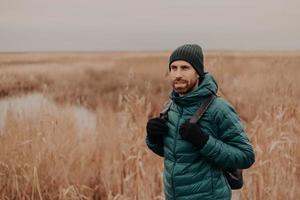 cara com a barba pensativa usa chapéu, jaqueta verde e luvas, carrega mochila, posa contra o fundo amarelo do campo de outono, passeia lá fora, respira ar fresco, focado na distância. conceito de turismo foto