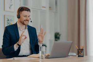 conceito de trabalho de carreira e distância. homem barbudo satisfeito trabalhador corporativo vestido formalmente gesticula bem sinal na webcam do laptop usa fone de ouvido para poses de chamadas on-line na área de trabalho ganha novos conhecimentos foto