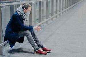 turista cansado faz uma pausa, usa celular, veste jaqueta, jeans e tênis, usa máscara de proteção contra coronavírus, se preocupa com a saúde em local público, lê informações. conceito de segurança foto