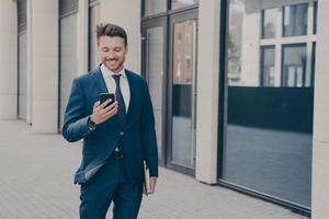consultor de negócios masculino jovem feliz confiante segurando smartphone moderno e sorrindo ao ar livre foto
