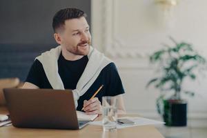 alegre estudante do sexo masculino procura informações para lição de casa via laptop escreve artigo senta-se no espaço de coworking sozinho desvia o olhar com sorriso feliz no rosto relógios treinamento webinar cria blog pessoal foto