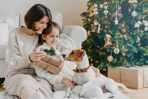 jovem feliz abraça sua filhinha, segura o presente de natal, aguarda as férias de inverno, brinca com o filhote de pedigree, posa no chão perto da árvore decorada do ano novo. conceito de tempo familiar feliz foto