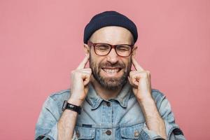 macho barbudo irritado tapa os ouvidos quando ouve um som desagradável, mantém os olhos fechados, insatisfeito com algo, isolado sobre fundo rosa. homem de meia idade não barbeado ignora ouvir alguém foto