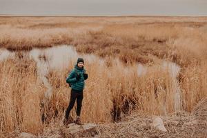 foto de homem ativo vestido com roupas da moda, fica na pedra contra perto de um pequeno lago e campo amarelo de outono, carrega mochila, copie o espaço de lado para seu conteúdo publicitário ou informações