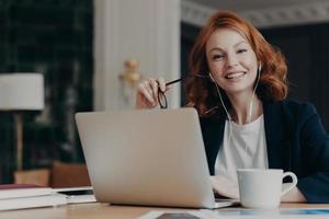 jovem freelancer feminina feliz com cabelo ruivo pensa em novas ideias para o trabalho, trabalha à distância no computador portátil, prepara-se para videoconferência, usa fones de ouvido, posa no local de trabalho com bebida quente foto