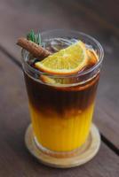 café preto gelado de close-up misturado com suco de laranja em cobertura de vidro com laranjas fatiadas, folhas de alecrim e canela por cima. em uma montanha-russa de madeira colocada sobre uma mesa de madeira. foto