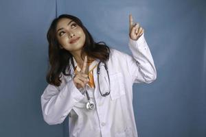 retrato de uma jovem médica asiática, um profissional médico está sorrindo e apontando para cima em um espaço de cópia isolado sobre fundo azul foto