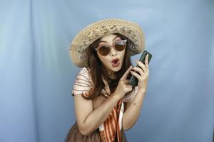 jovem mulher asiática apontando e chocando seu smartphone isolado por um fundo azul foto