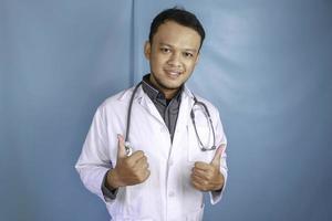 retrato de um médico jovem asiático, um profissional médico está sorrindo e mostrando os polegares para cima ou sinal de ok isolado sobre fundo azul foto