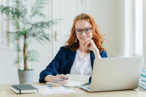 foto de freelancer ruiva de sucesso tem trabalho remoto, assiste webinar on-line no computador portátil, anota informações no bloco de notas, sorri alegremente posa no local de trabalho vestido com roupas formais