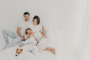 pose de família feliz na cama branca durante o fim de semana. pai, mãe, filha e cachorro sentem o conforto de estar no quarto, abraçam e se divertem, estando relaxados e alegres. família amigável e descanso foto