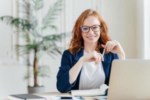 retrato de empregado de mulher ruiva feliz em óculos ópticos, tem expressão satisfeita, trabalha com aparelhos modernos, aguarda a reunião com o colega, prepara o relatório contábil, senta-se no próprio gabinete foto