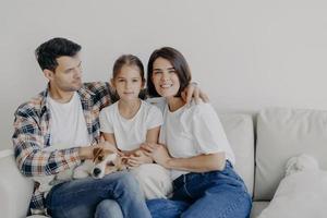 conceito de tempo em família. alegre mãe, pai e garotinha abraçam-se todos juntos, passam o tempo livre na sala de sua nova casa, sentam-se no sofá, brincam com o cachorro, têm relacionamentos perfeitos, desfrutam de conforto foto