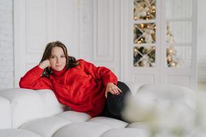 foto de jovem relaxada vestida com roupas de malha quentes, encontra-se no sofá confortável, goza de uma atmosfera doméstica, árvore de ano novo decorada no fundo. conceito de pessoas, lazer e tempo livre