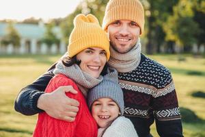 adorável criança fica perto de seus pais afetuosos, gosta de passar tempo juntos, abraçam-se, sorriem alegremente, olham diretamente para a câmera. agradáveis momentos inesquecíveis com a família foto