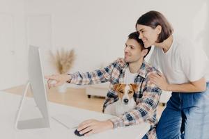 marido feliz aponta para o monitor do computador, pede conselhos úteis à esposa o que é melhor comprar, faz compras on-line, posa na área de trabalho com o animal de estimação favorito. pessoas, família e conceito de tecnologia foto