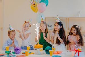 crianças e conceito de férias. feliz por cinco amigos olharem alegremente no bolo com brilho, comemorar aniversário, usar chapéus de cone de festa e segurar balões de ar, ter expressões felizes foto