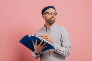 tiro de homem com barba pensativo concentrado de lado, pensa sobre o que escrever. arquiteto masculino bonito detém livro e lápis de cor, sublinha as informações para o projeto, isolado sobre fundo rosa. foto