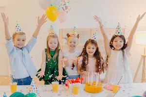 tiro horizontal de crianças felizes e positivas pegam confete, comemoram aniversário juntos, levantam os braços, têm bom humor, brincam juntos, ficam perto de mesa festiva com caixa de presente, bolo, chapéus de festa