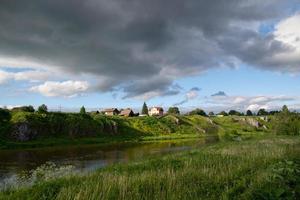 uma vila na margem do rio com grama verde brilhante e um lindo céu. foto