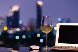 um copo com vinho branco coloca na mesa com computador portátil e luzes coloridas de bokeh da cidade. trabalhando em casa conceito. foto