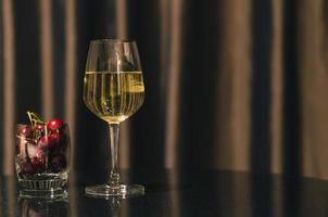um copo de vinho branco com frutas cereja colocado na mesa na sala com fundo de cortina. foto