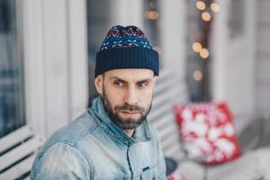 homem europeu bonito com barba grossa e bigode usa chapéu e jaqueta jeans, tem expressão pensativa, contempla algo, posa no interior. homem elegante barbudo jovem pensativo. foto