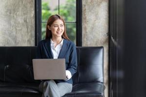 mulher asiática digitando e-mail no computador portátil enquanto está sentado em casa, jovem empresária trabalhando no escritório moderno de inicialização criativa. analisar ações de mercado, nova estratégia. foto