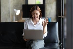 jovem asiática tem bate-papo por vídeo com amigos com webcam, feliz jovem empresária asiática acenando com as mãos para cumprimentar o parceiro durante a videoconferência com sua equipe. foto