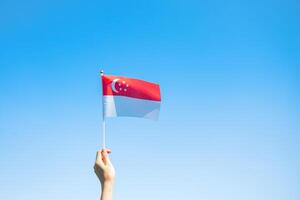 mão segurando a bandeira de Cingapura no fundo do céu azul. dia nacional de singapura e conceitos de celebração feliz foto