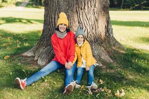 beautiul feminino usa chapéu de malha e swetaer senta-se junto com sua filha perto da árvore, descansa ao ar livre no belo parque, desfruta do ar fresco do outono, sente-se relaxado. pessoas, relaxamento foto