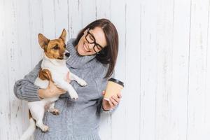 modelo feminino morena feliz usa suéter de malha quente, óculos, carrega seu cachorro favorito e café para viagem, vai passear juntos, ter bons relacionamentos amigáveis. conceito de pessoas e animais de estimação foto