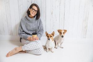 foto interna de mulher bonita usa óculos e roupas casuais, bebe bebida quente, gosta de bom dia, senta-se perto de seus animais de estimação favoritos contra o fundo da cerca branca. conceito de felicidade e lazer