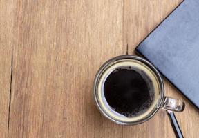 vista superior do café americano quente preto em uma xícara em uma mesa de madeira rústica com um livro e lápis. relaxar e o conceito de pausa para o café foto