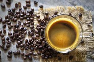 xícara de café americano preto quente em uma mesa de concreto rústica de pano de saco com uma pilha de grãos de café arábica marrom torrado. atualizar, conceito de bebida matinal foto