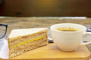 xícara de café preto em uma placa de madeira com sanduíche de presunto e queijo em uma mesa de concreto rústica foto