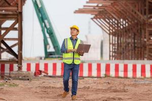 engenheiro verificando projeto no canteiro de obras, homem de capacete com laptop no canteiro de obras de infraestrutura foto