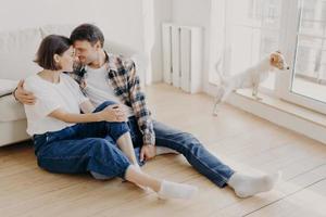 tiro horizontal de marido e mulher satisfeitos abraçam e beijam, têm um bom relacionamento, sentam-se no chão de madeira perto do sofá, gostam de estar em casa durante o fim de semana, moram em apartamento novo, começam uma nova vida em residência foto