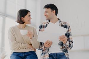 foto de marido e mulher caucasianos felizes discutem a compra de um novo apartamento, o homem detém documentos, sugere alugar um novo apartamento, a mulher bebe café da xícara, posam juntos no interior, estando em casa