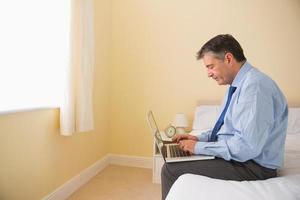 homem concentrado usando seu laptop sentado em uma cama foto