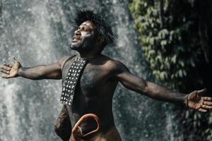 close-up de papua homem da tribo dani estende as mãos sente liberdade, contra cachoeira na floresta verde foto