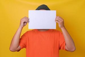cara jovem de camiseta laranja, escondendo o rosto sob papel branco lblank, fundo amarelo isolado. horizontal, papel estilo paisagem. foto