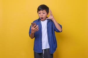 menino asiático em estilo jeans casual está segurando o telefone e mostrando expressão de surpresa isolada em fundo amarelo. foto