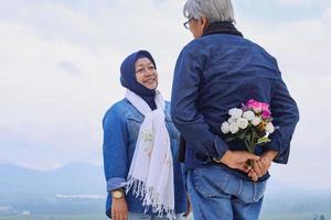 verso do velho escondendo flor enquanto enfrenta sua esposa sorrindo contra a paisagem dramática. vista da plantação de chá, montanha e lago. casal de idosos românticos. foto