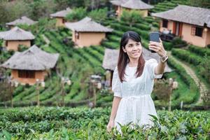 mulher de turista feliz no vestido branco tirando foto pelo smartphone móvel no lindo chá garden.traveler visitando na vila tailandesa de ban rak, mae hong son, tailândia. conceito de viagens, férias e férias